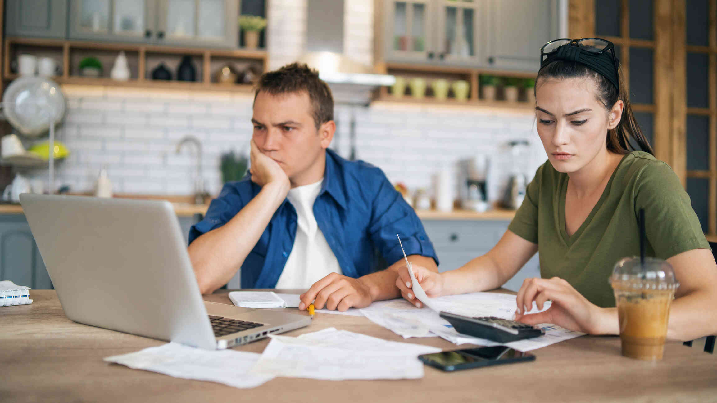 A couple is seated at a kitchen table, the woman looking through the documents while the man is busy on the laptop.